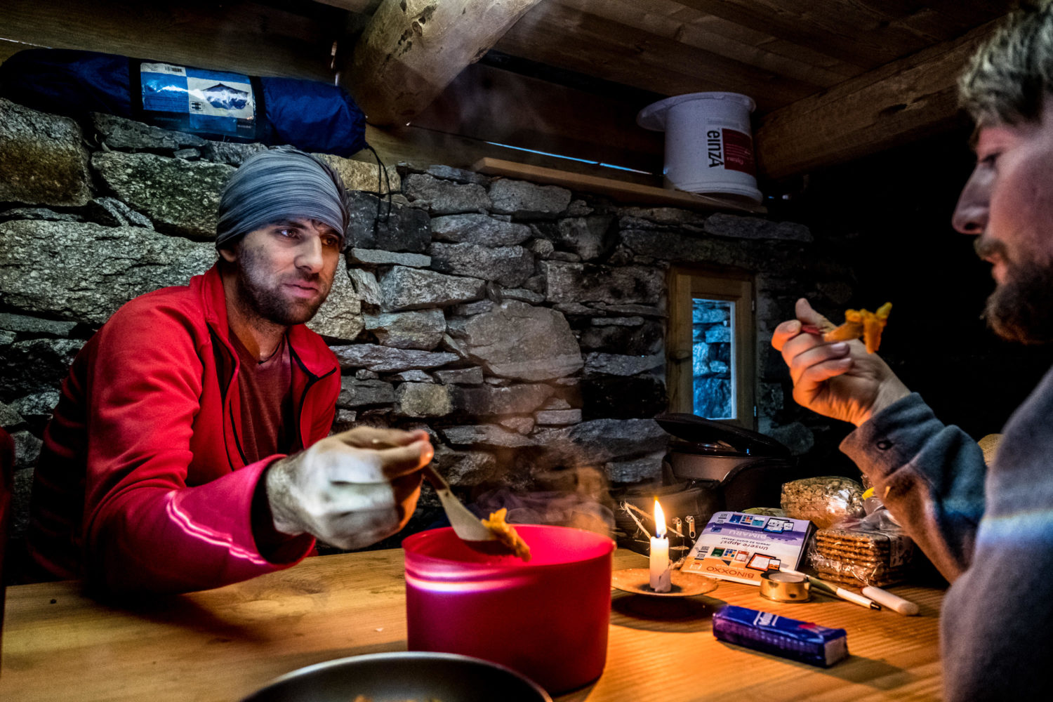 Zwei Wanderer essen Nudeln aus einem Topf in einer Steinhütte bei Kerzenlicht